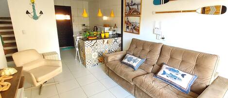 A living room with a brown sofa, a beige armchair, and a coffee table. There is a kitchen area with a tiled backsplash and yellow pendant lights.