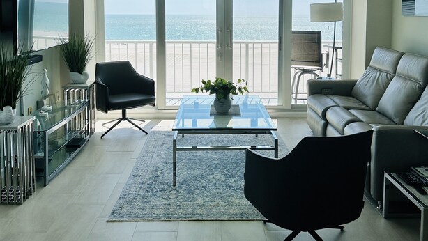 A spacious living room with a large window overlooking the ocean, a ceiling fan, and a modern sofa. - Wake up to the sound of waves and panoramic Gulf views at this beachfront condo  (Marco Island)