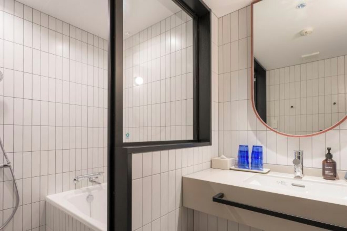 Un baño moderno con paredes de azulejos blancos, un espejo grande, una bañera y un mueble de tocador con lavabo y artículos de aseo.