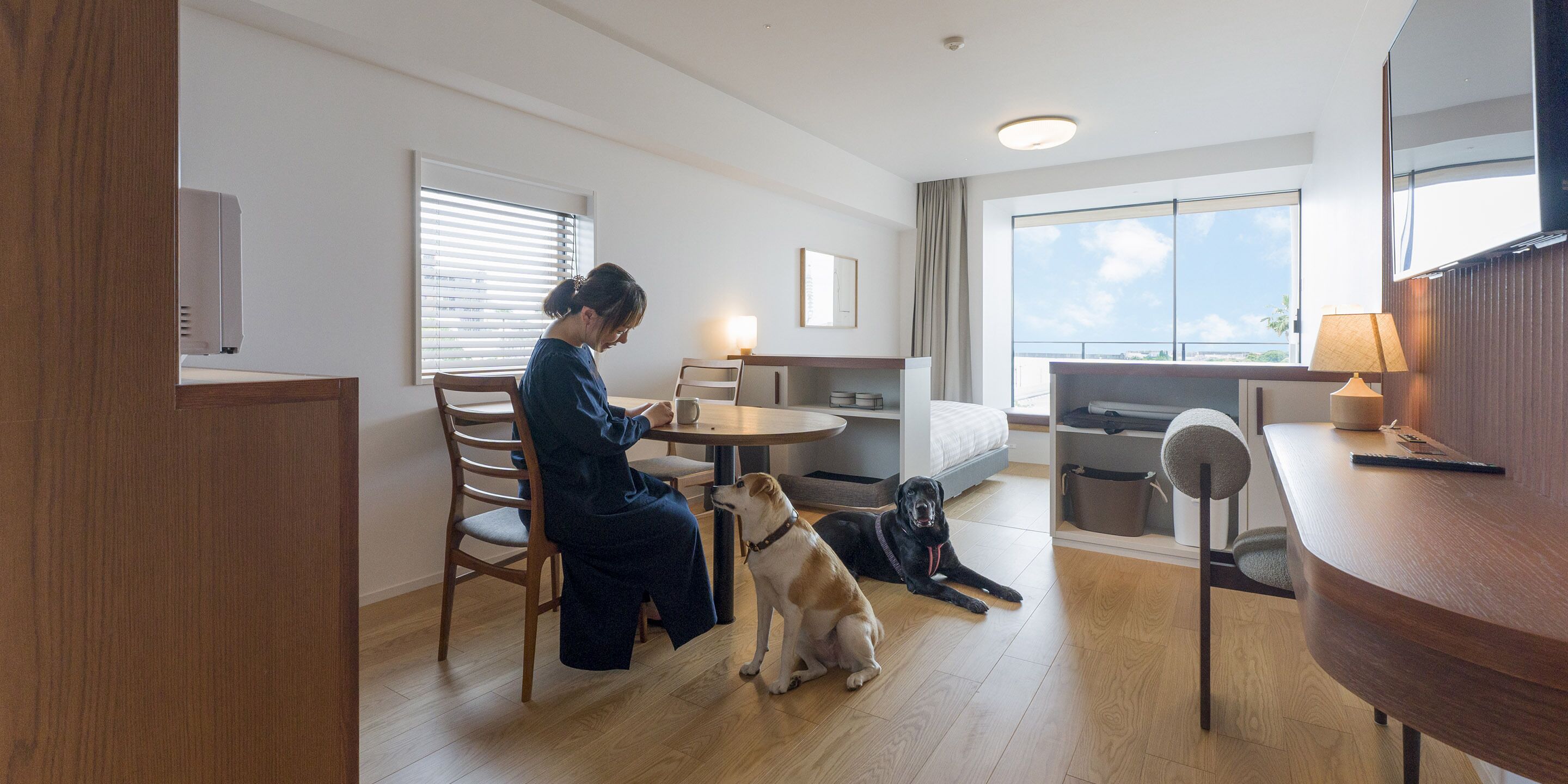 A modern hotel room with a wooden floor, a dining area with a table and chairs, a sofa, a large window with a view, and a desk with a lamp.