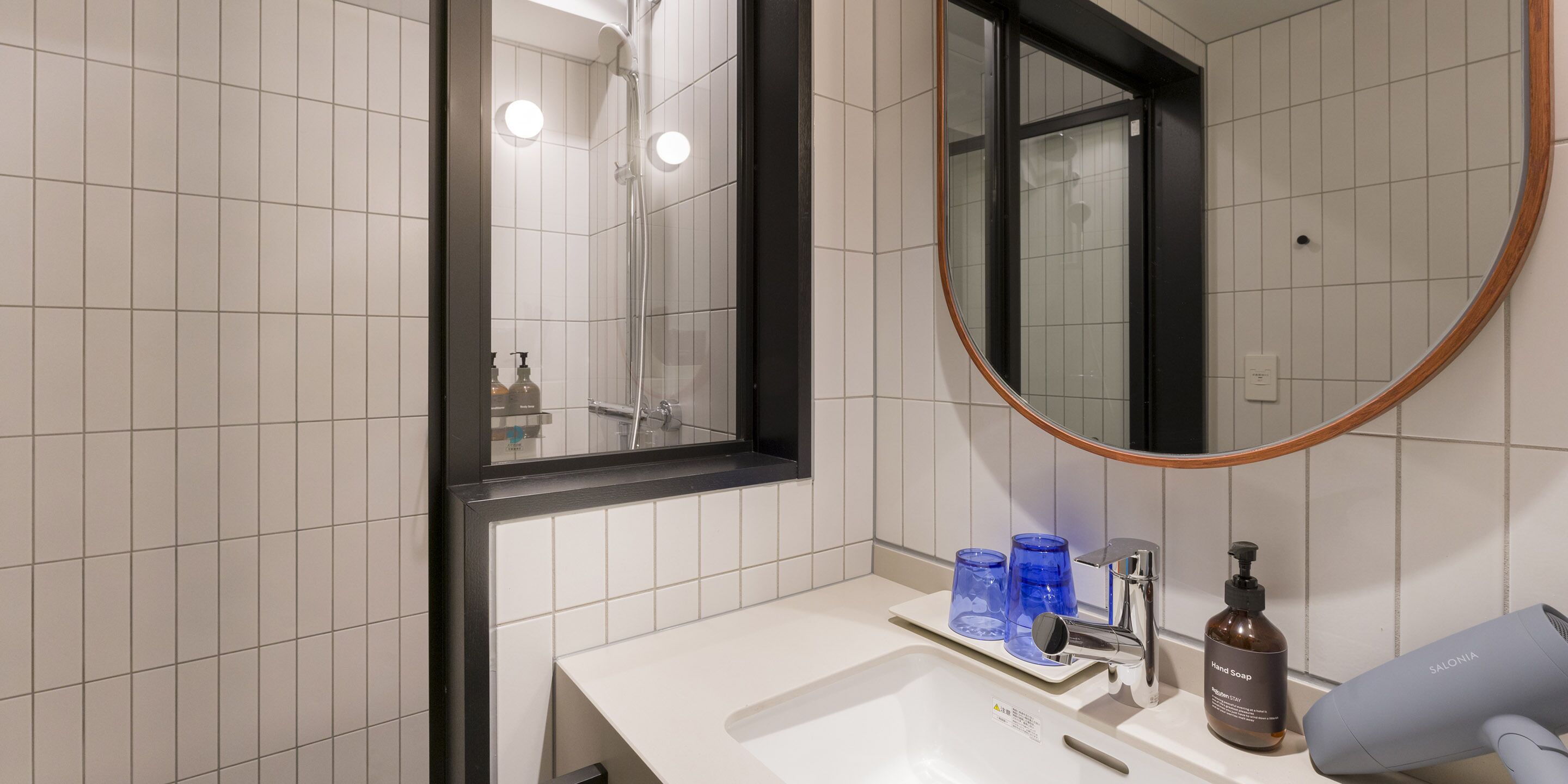 A bathroom with a white sink, a mirror, and a shower. There are blue glasses on the sink and a hairdryer on the counter.