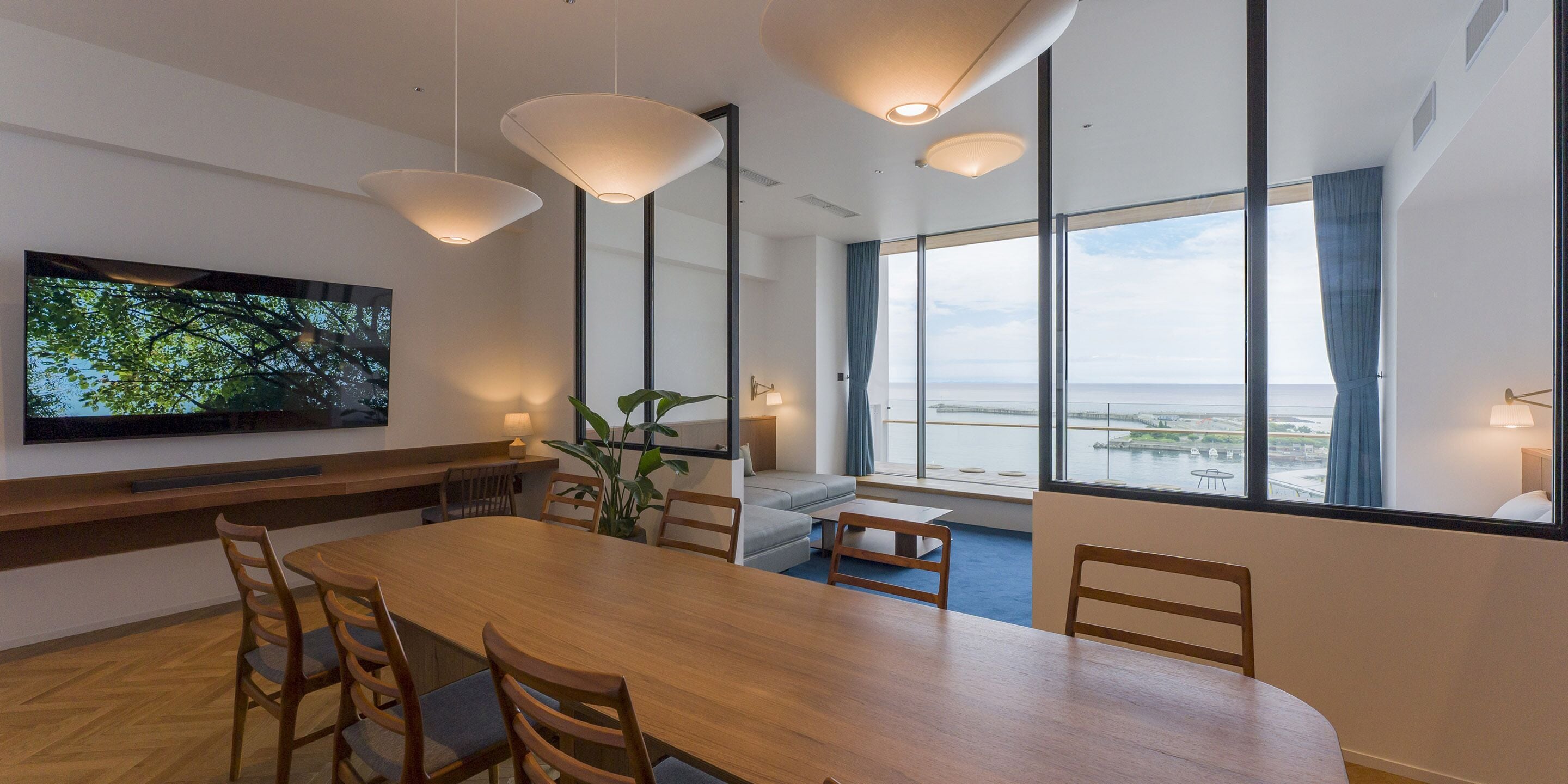 A modern dining area with a large wooden table, multiple chairs, pendant lights, a television on the wall, and large windows offering a view of the sea.