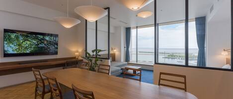 A modern dining area with a large wooden table, multiple chairs, pendant lights, a television on the wall, and large windows offering a view of the sea.
