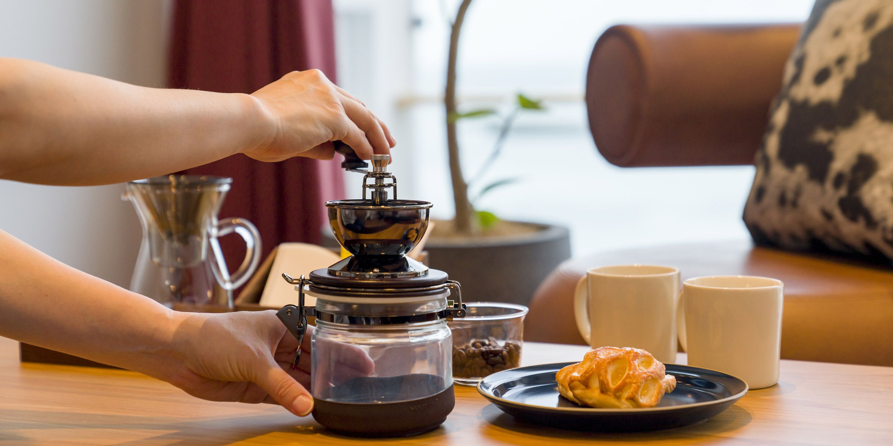 A person using a coffee grinder, a plate with a pastry, and two coffee cups on a table.