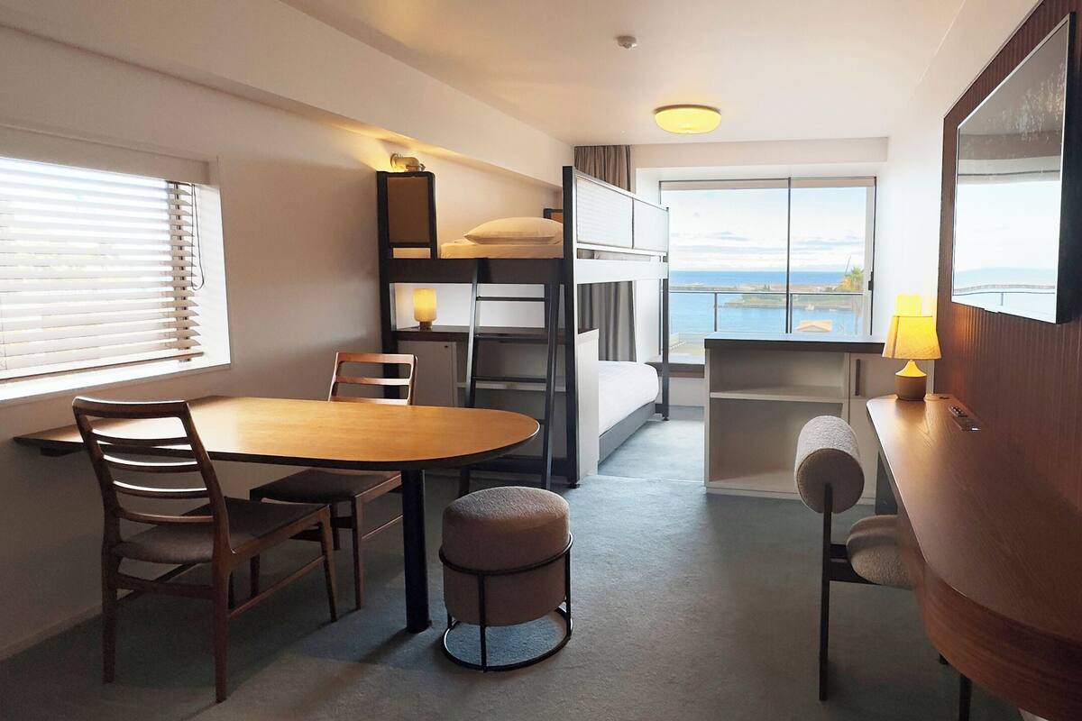 Habitació d'hotel petita amb lliteres, zona de menjador amb taula i cadires, i una finestra gran que ofereix vista al mar.