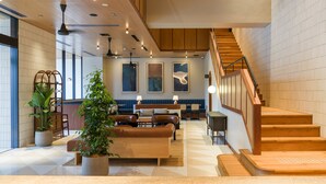 Eine moderne Hotellobby mit einer Holztreppe, einer Sitzecke mit Sofas und einem Couchtisch sowie einer großen Topfpflanze.