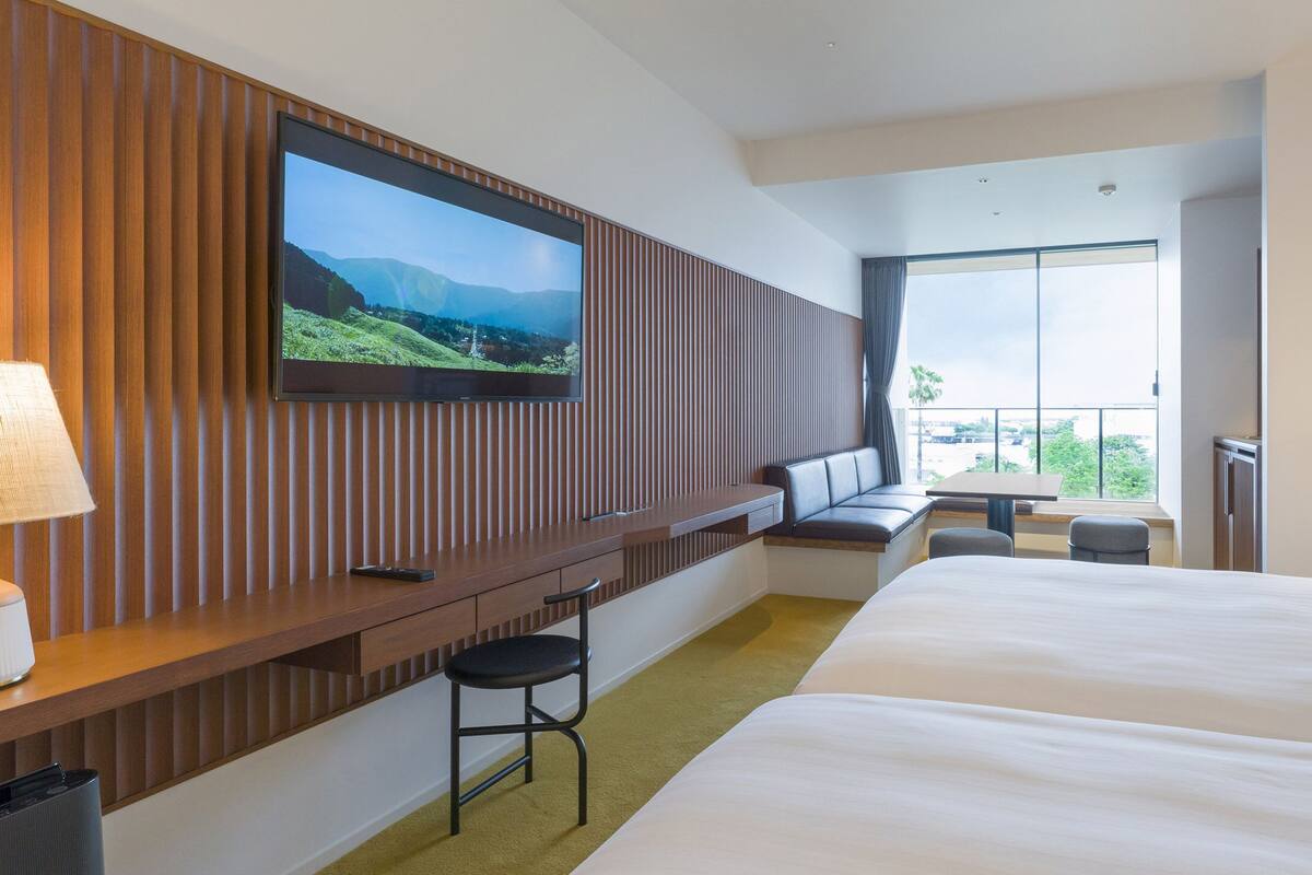 Habitació d'hotel amb un llit gran, un televisor muntat a una paret de panells de fusta, una zona de seients amb una taula i cadires, i una finestra gran que ofereix vistes a l'exterior.