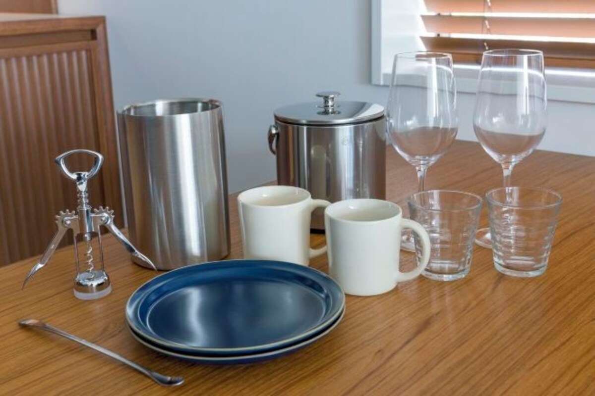 Una mesa de madera con un plato azul, dos tazas blancas, una cafetera de metal, un recipiente metálico, un destapador de vino y dos copas de vino.