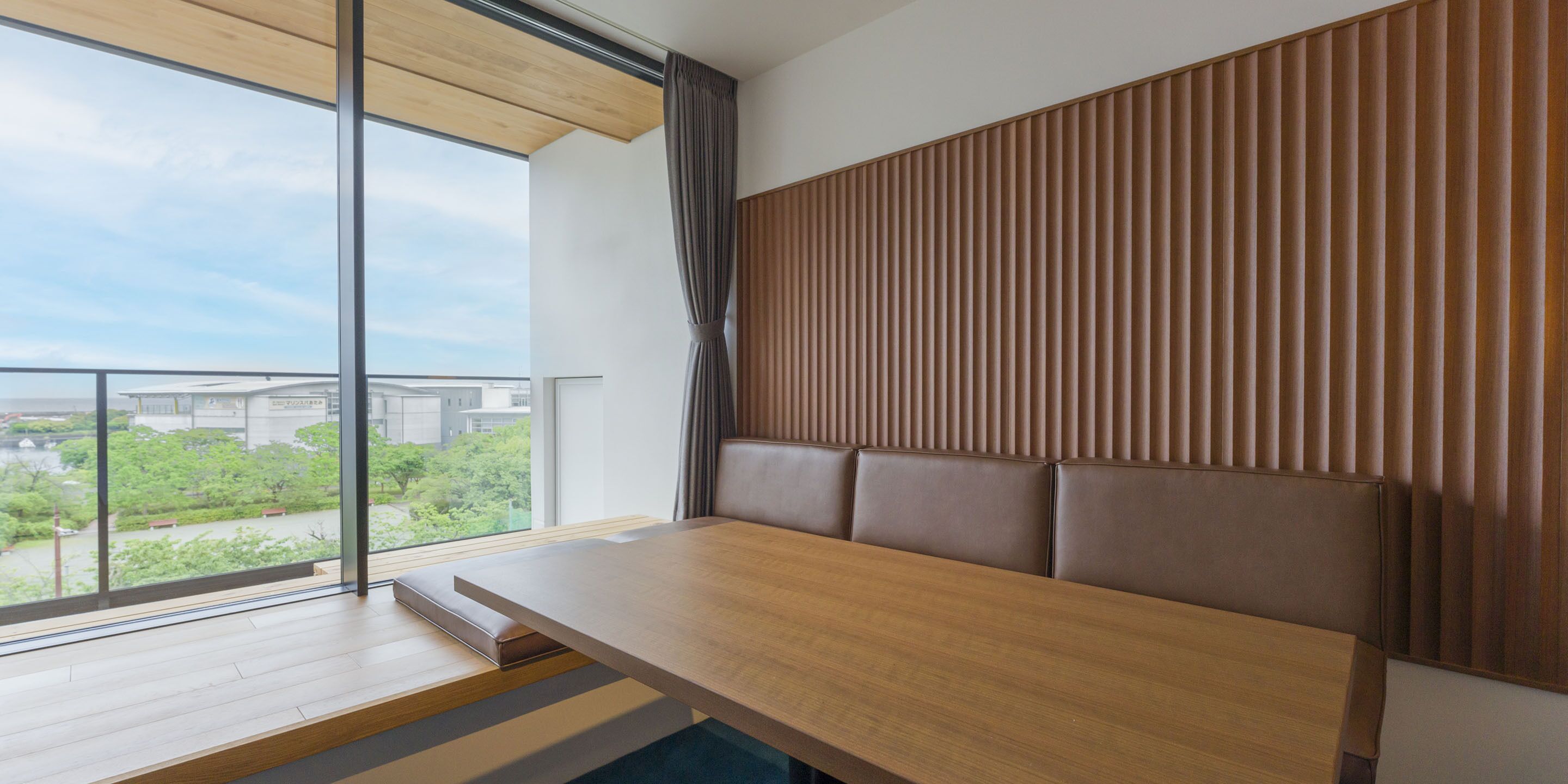 A modern room with a wooden table and bench, large windows with a view of greenery and buildings, and a wooden slatted wall.