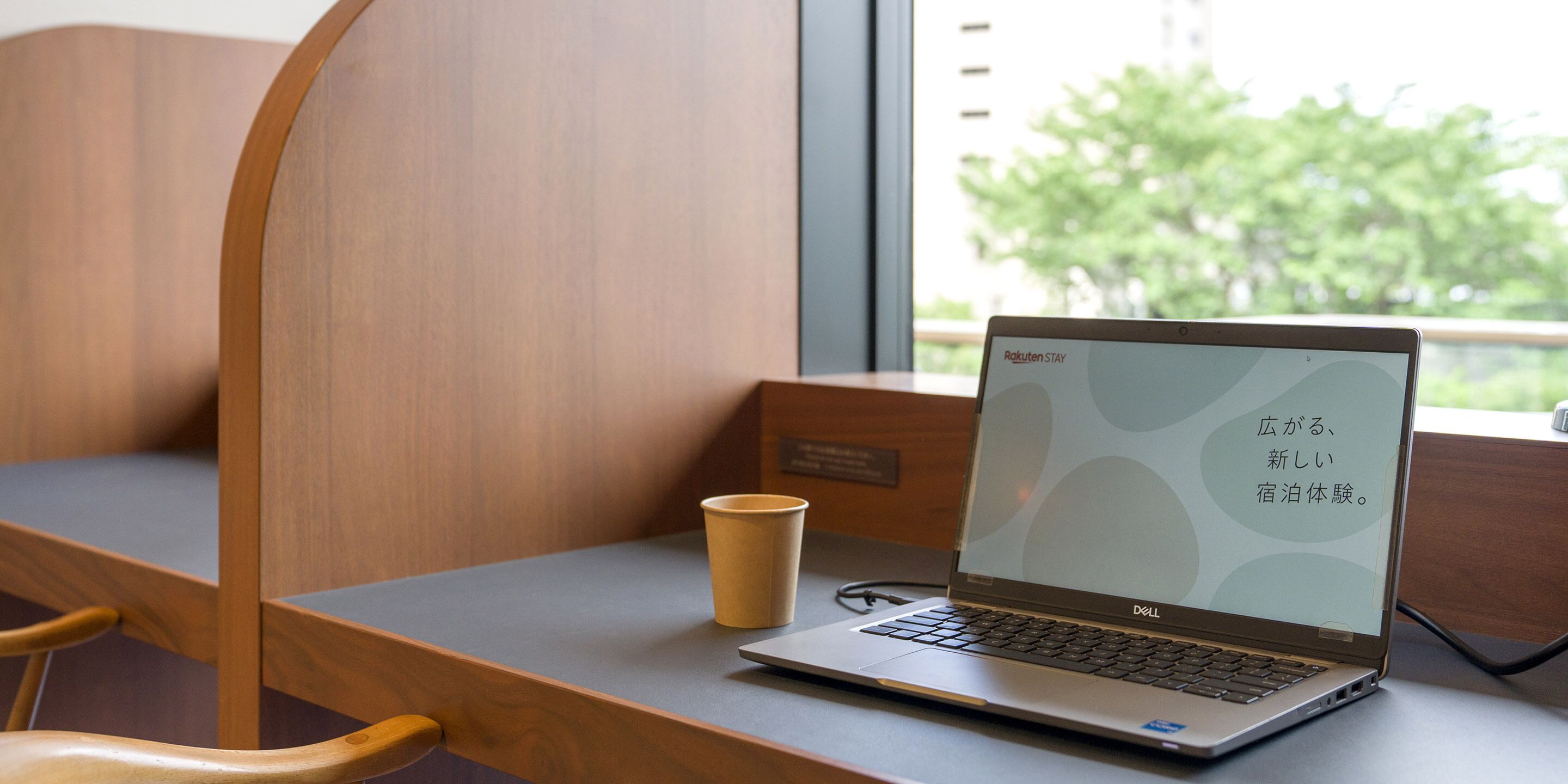 A laptop on a desk with a cup beside it, a window with a view of greenery, and a wooden chair.