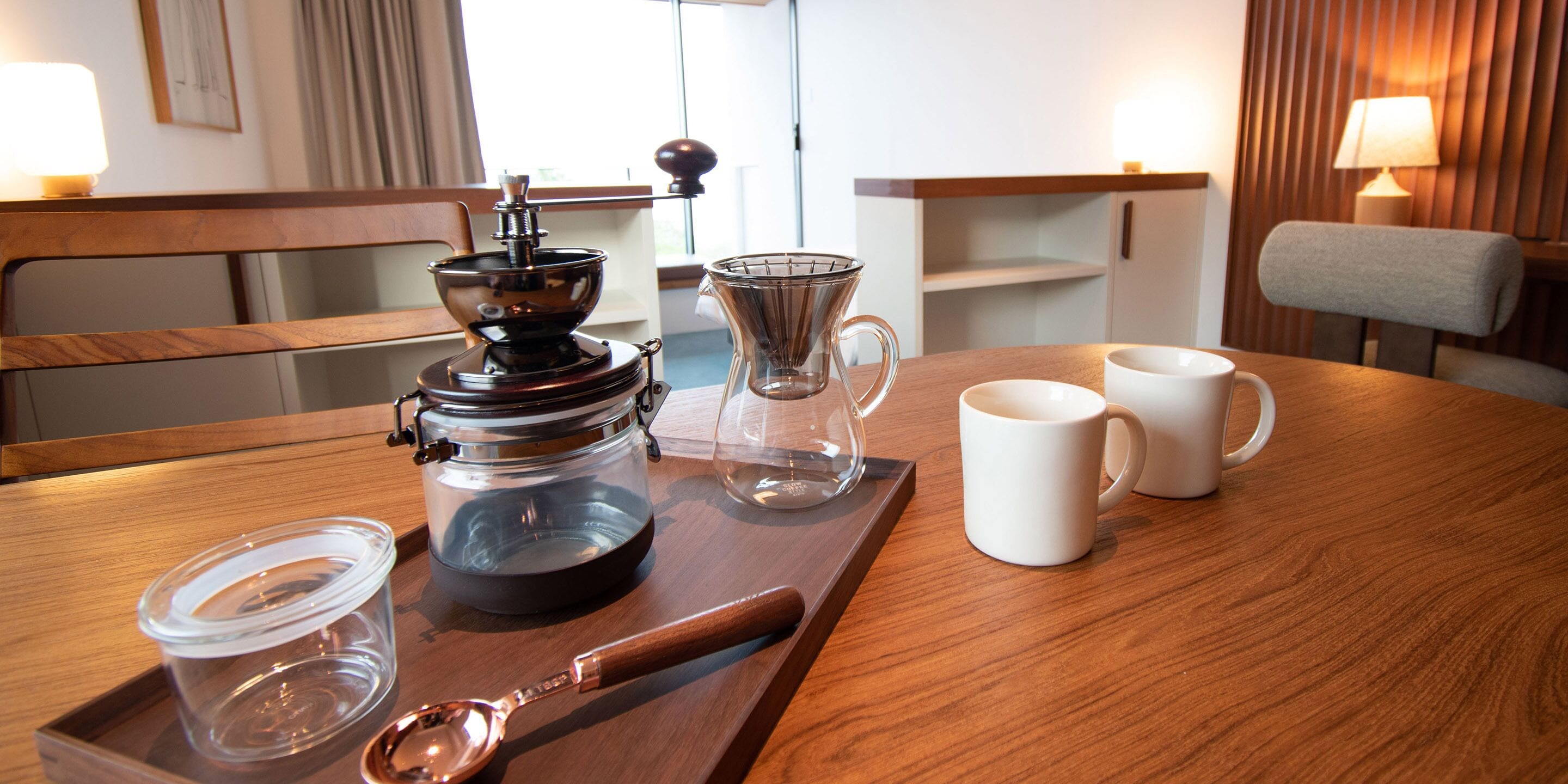 A wooden table with a coffee maker, a glass carafe, two white mugs, a spoon, and a small container.