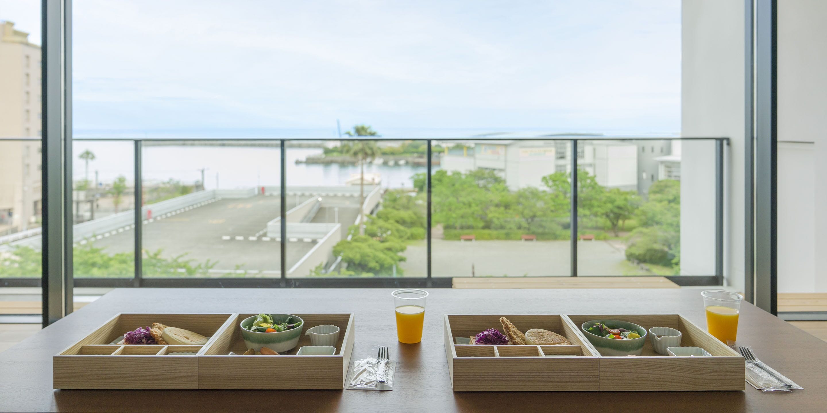Una preparación para el desayuno con dos bandejas de madera, cada una con un plato de comida, un vaso de zumo de naranja y una taza pequeña. La habitación tiene ventanas grandes con vistas a un puente y follaje.