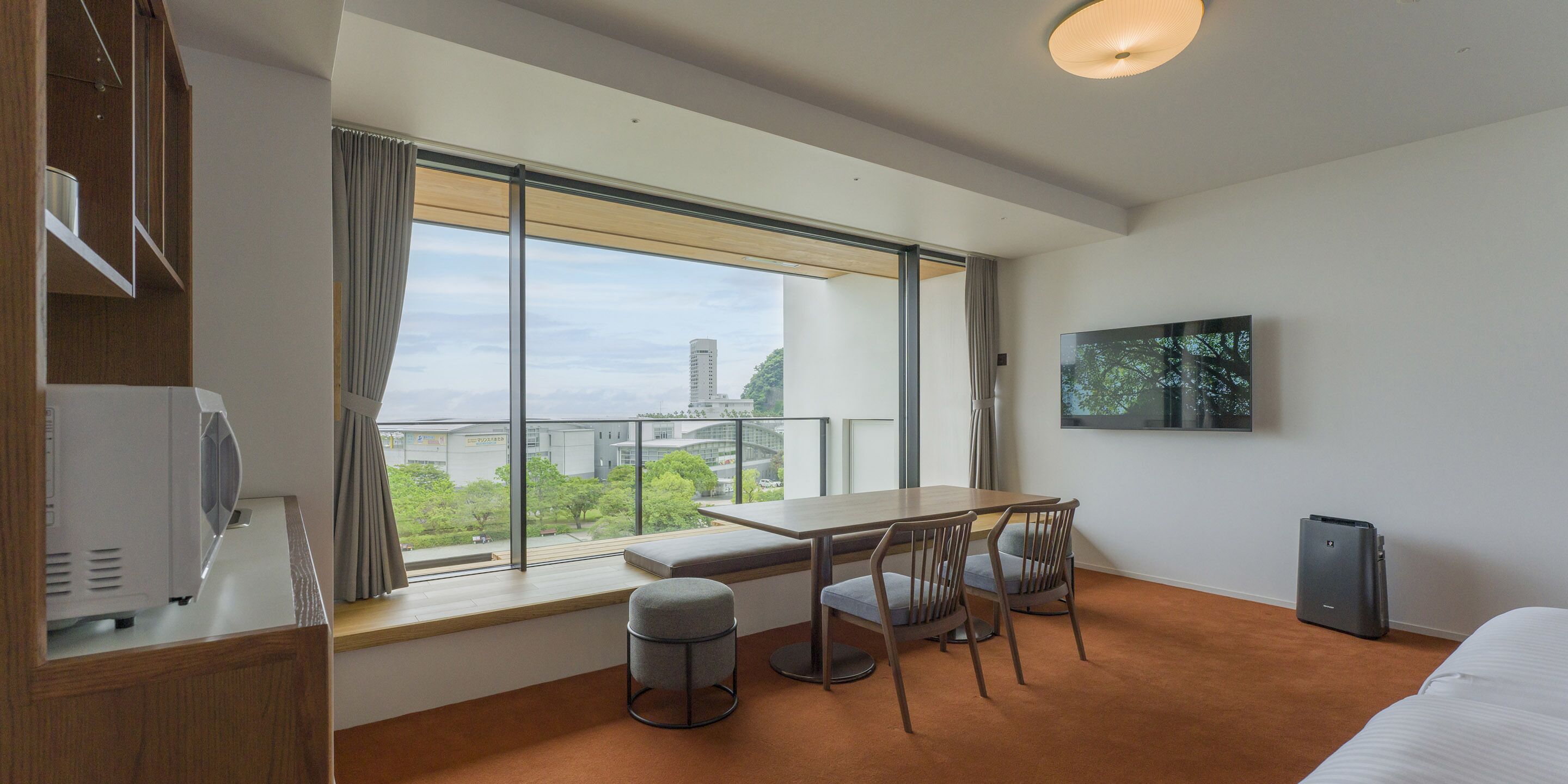 Una habitación de hotel moderna con un ventanal grande, una mesa de comedor con sillas, un banco, un televisor y un mueble pequeño.