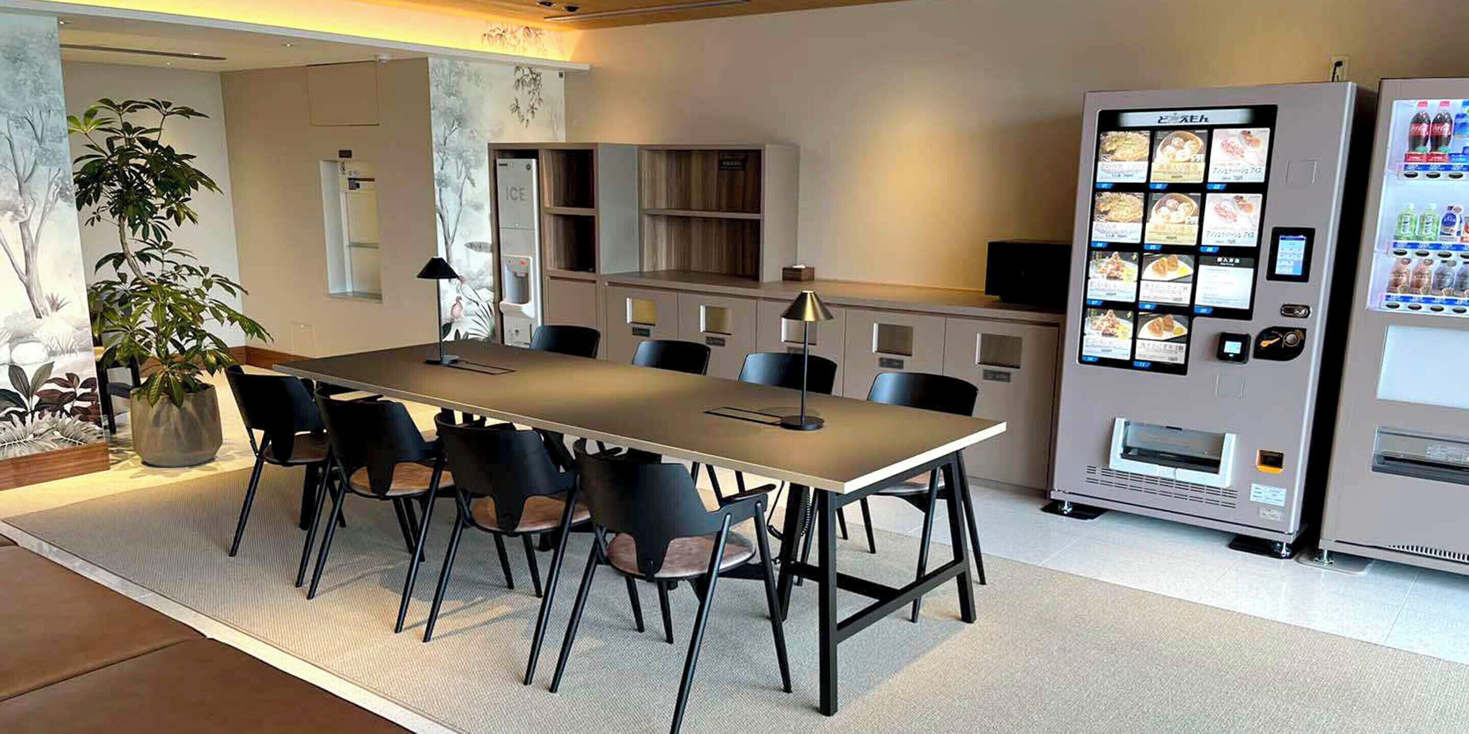 A modern dining area with a long table and chairs, a vending machine, and a refrigerator.