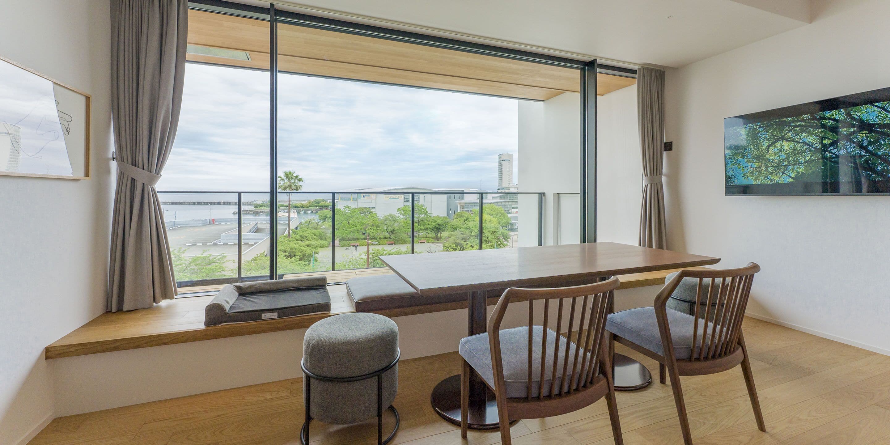 Une chambre moderne avec une grande fenêtre, une table en bois, deux chaises et un petit tabouret.