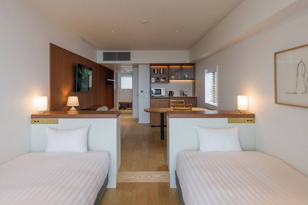 Habitació d'hotel amb dos llits individuals, una cuineta petita, un televisor fixat a la paret i una obra d'art emmarcada sobre el llit.