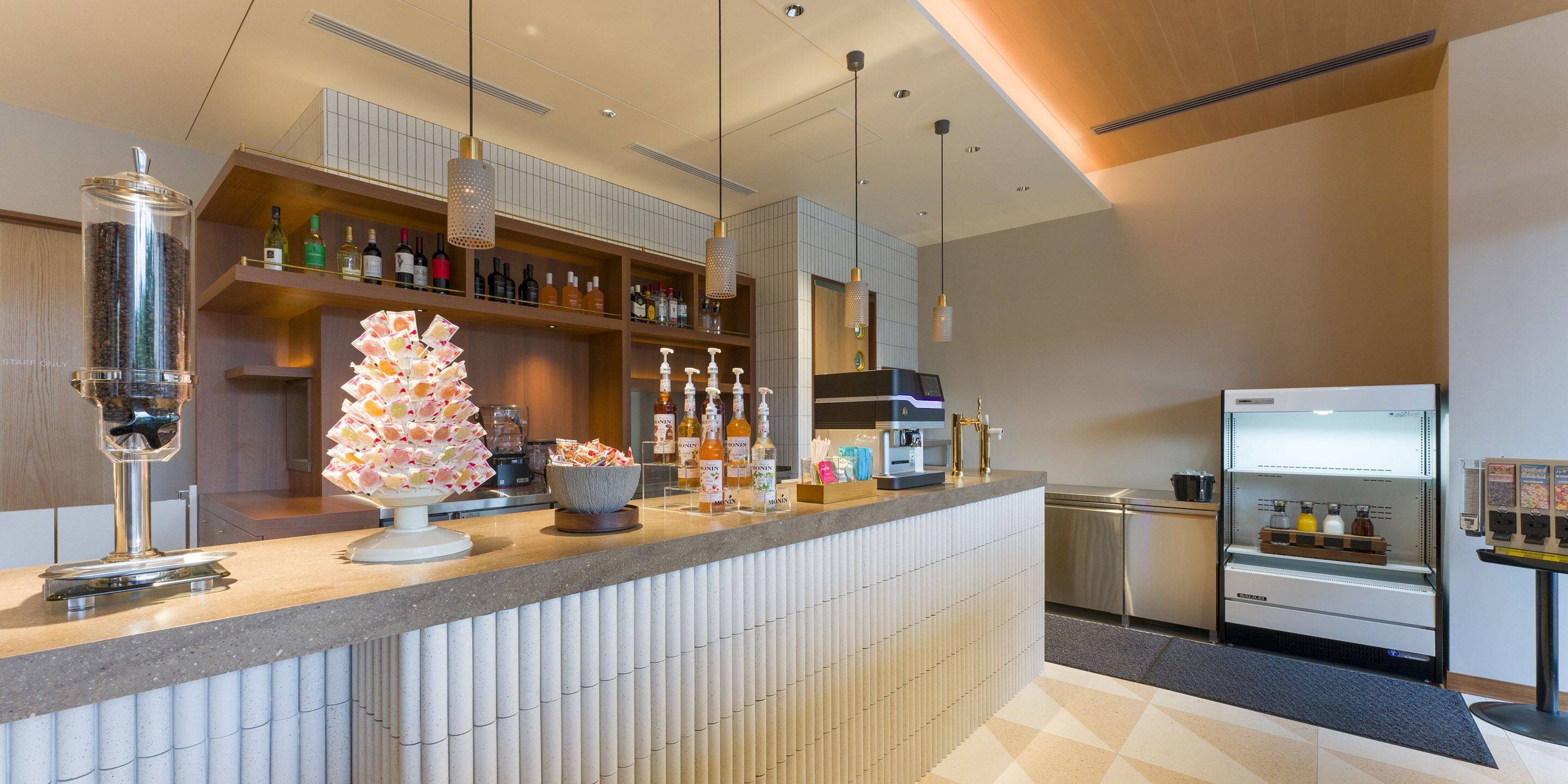 Eine moderne Bar mit Theke, Regalen voller Flaschen, einer Kaffeemaschine und einem Kühlschrank.