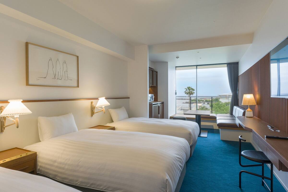 Habitació d'hotel amb dos llits, un escriptori, una cadira, una taula i una finestra gran que ofereix una vista.
