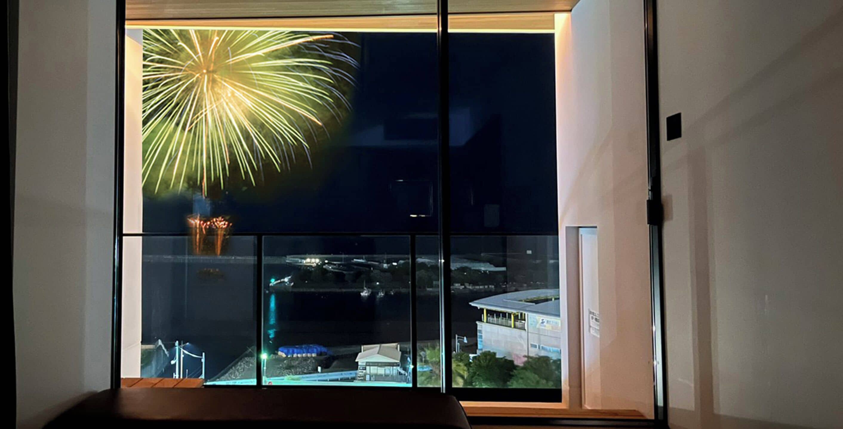 Una habitación con un ventanal grande que ofrece vistas a un paisaje urbano nocturno, con un espectáculo de fuegos artificiales.