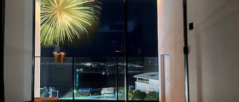 A room with a large window overlooking a cityscape at night, featuring a fireworks display.