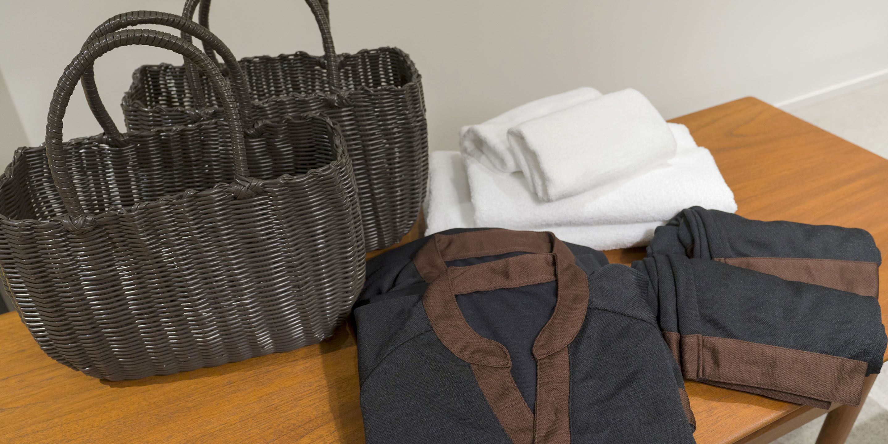 A wicker basket, folded white towels, and a dark shirt with brown trim on a wooden table.