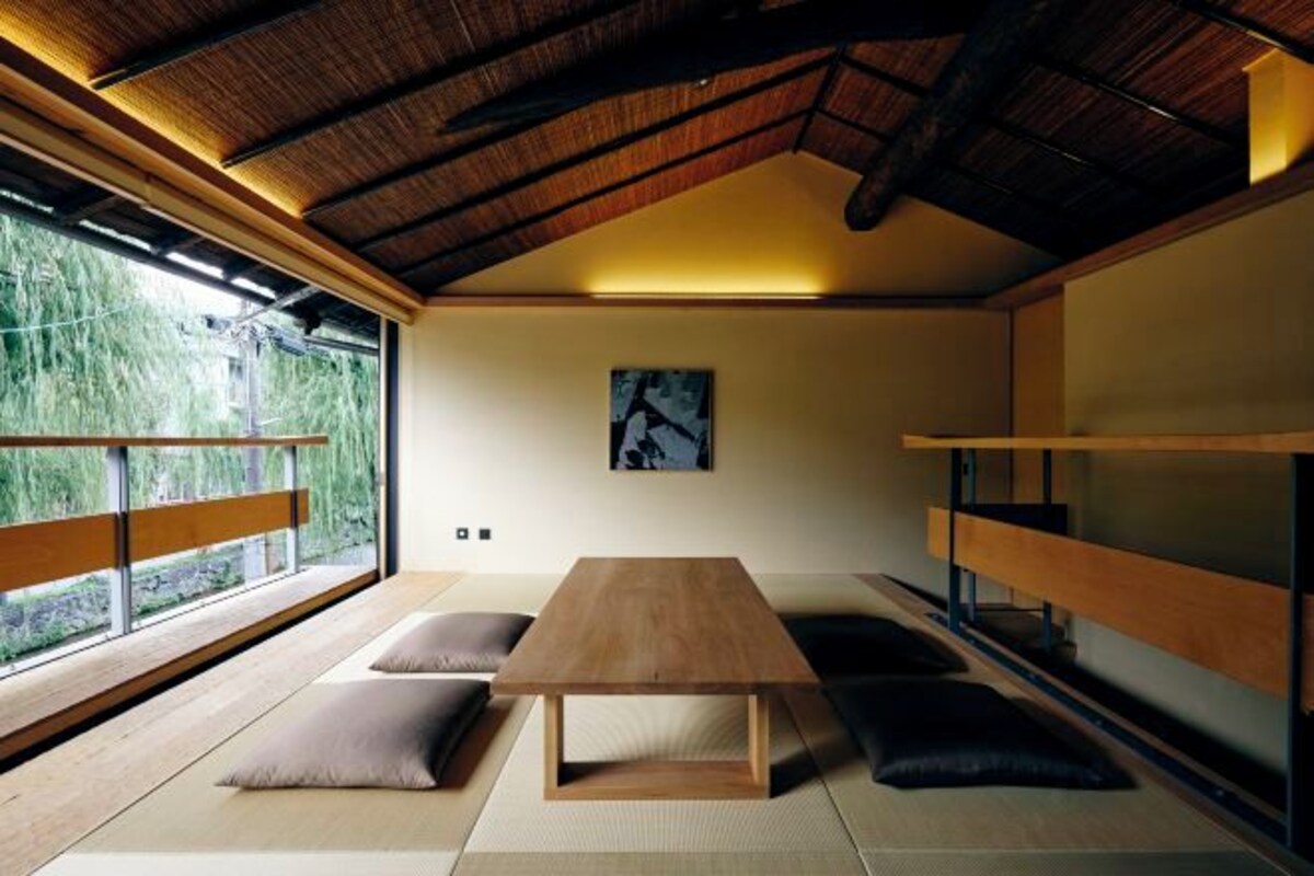 Une pièce minimaliste avec un sol recouvert de tatamis, une table en bois, des coussins, une grande fenêtre donnant sur un paysage verdoyant et un plafond avec des poutres apparentes.
