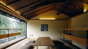 Ein Zimmer mit Tatami-Boden, einem Holztisch, Kissen, einem Etagenbett und einem großen Fenster mit Blick auf Grünflächen.