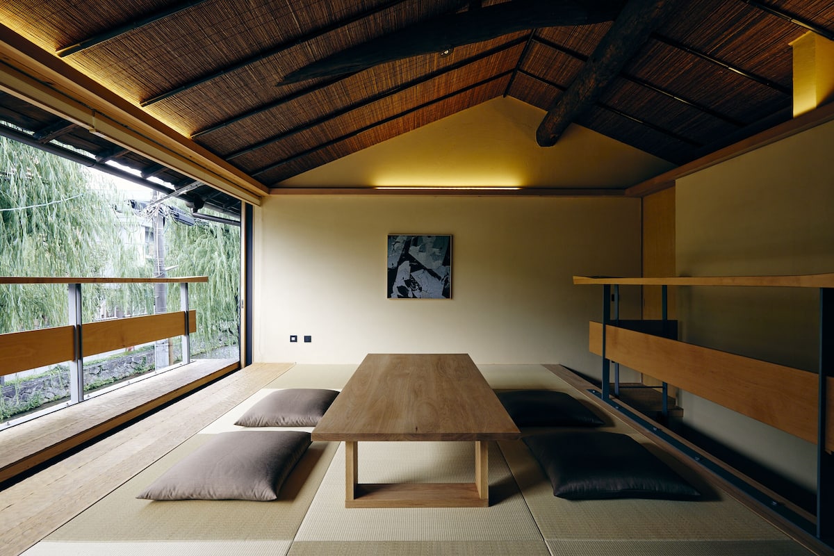 Quarto com piso de tatame, mesa de madeira, almofadas, beliche e uma janela grande com vista para o verde.