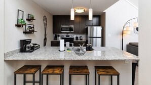 A modern kitchen with dark cabinets, stainless steel appliances, a granite countertop, and bar stools. - Downtown Loft Skyline View 2BR (Des Moines)