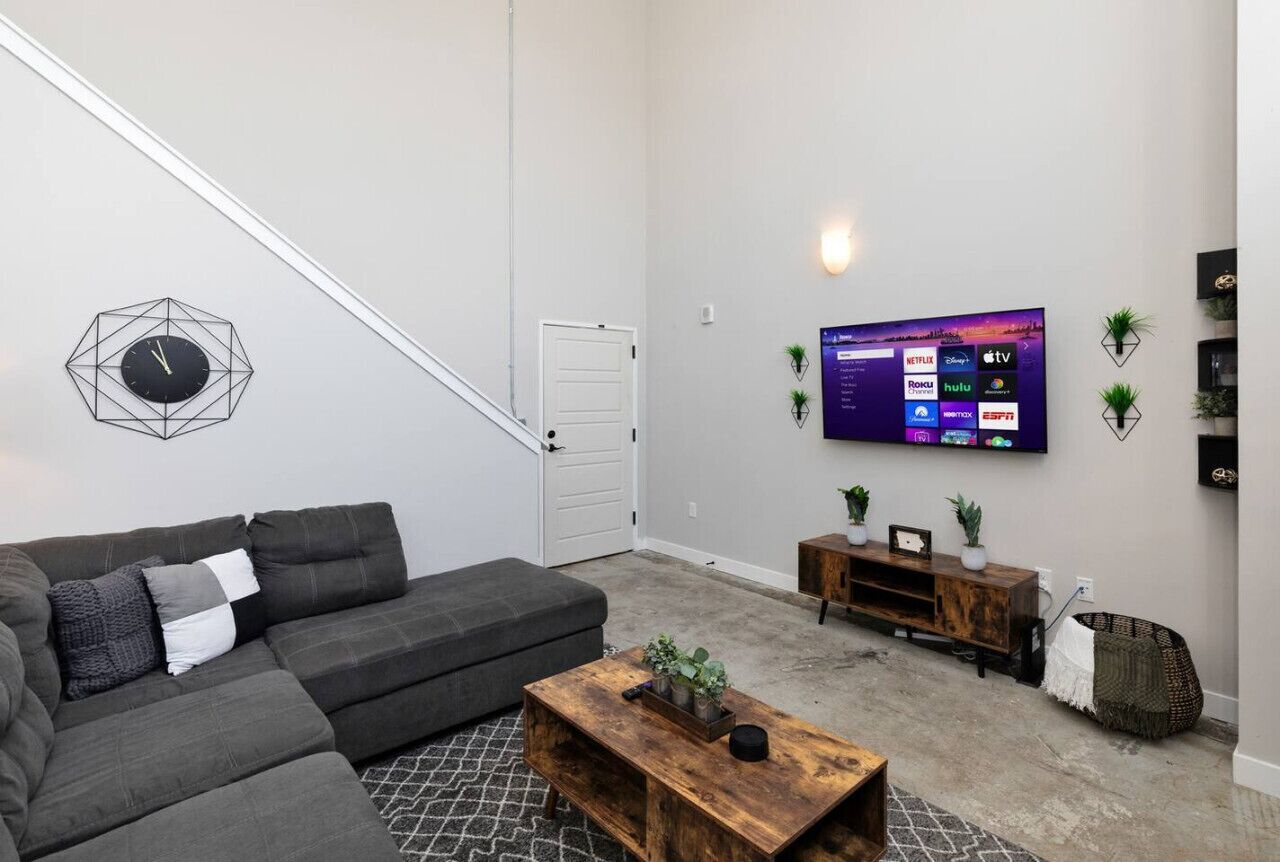 Uma sala de estar moderna com um sofá seccional, uma mesa de centro de madeira, uma TV montada na parede e uma escada que leva a uma porta.