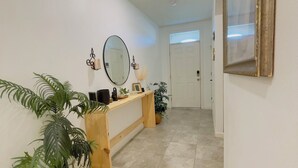 Um corredor com uma mesa consola de madeira, plantas em vasos, um espelho grande e uma porta branca no final.