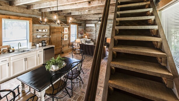Un interior rústico con vigas de madera a la vista, una escalera de madera, un comedor con mesa y sillas negras, y una cocina con armarios blancos y un ventanal.