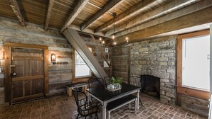 Un interior rústico con vigas de madera a la vista, una chimenea de piedra, una mesa de comedor con sillas y una escalera.