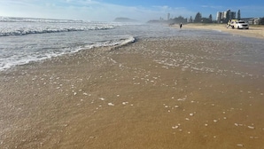 A beach with a sandy shore, waves, and a few people walking. There are buildings and trees in the background.