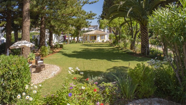 A garden with a lawn, trees, and a building with a tiled roof. - Charming Studio with Pool in Praia da Luz (Luz)