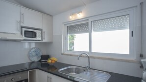 A modern kitchen with white cabinets, a black countertop, a stainless steel sink, a microwave, a toaster, and a window with blinds. - Charming Studio with Pool in Praia da Luz (Luz)