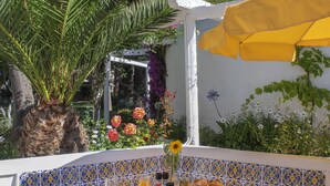 An outdoor dining area with a table set for breakfast, surrounded by plants and a white wall with blue and yellow tiles. - Charming Studio with Pool in Praia da Luz (Luz)