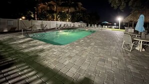 En udendørs swimmingpool med flisebelagt område, liggestole og et overdækket siddeområde om natten.