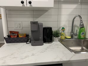 A kitchen with a coffee maker, a black electric kettle, a sink, and a plant.