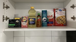 Een keukenkastje met diverse etenswaren, waaronder een doos Melange Irlandais, Lipton thee, zonnebloemolie van Plochman's, Atlantic zeezout, Redpath kristalsuiker en een pak bladerdeeg.