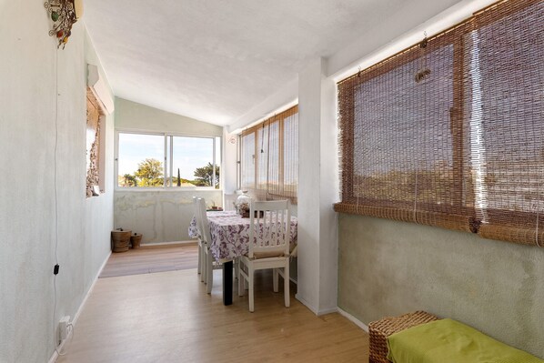 A room with a dining table, chairs, a green sofa, a window with a woven blind, and a view of the outdoors.