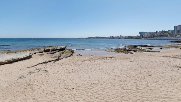 Una playa con costa rocosa, cielo azul claro y edificios al fondo.