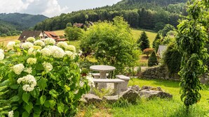 Une table et des bancs en pierre se dressent dans un jardin luxuriant, agrémenté de fleurs d’hortensia blanches, et sont entourés de verdure. En arrière-plan, on aperçoit un paysage rural avec des maisons et des collines.