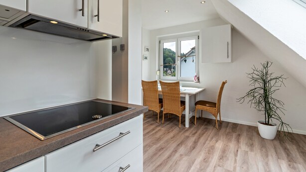 Une cuisine moderne dotée d’une cuisinière intégrée, de meubles blancs et d’un coin repas avec des chaises en osier et une plante en pot.
