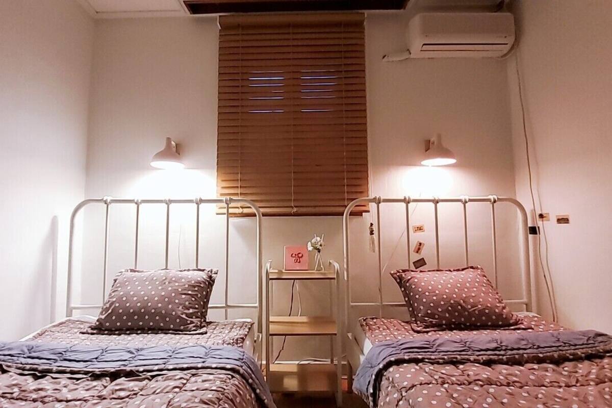 A twin bedroom with two beds, each with a metal frame and a pillow. There is a small table with a vase and a card between the beds.