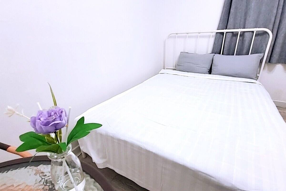A single bed with a white mattress and grey pillows, a small vase with a purple flower, and a round table with a white cloth.