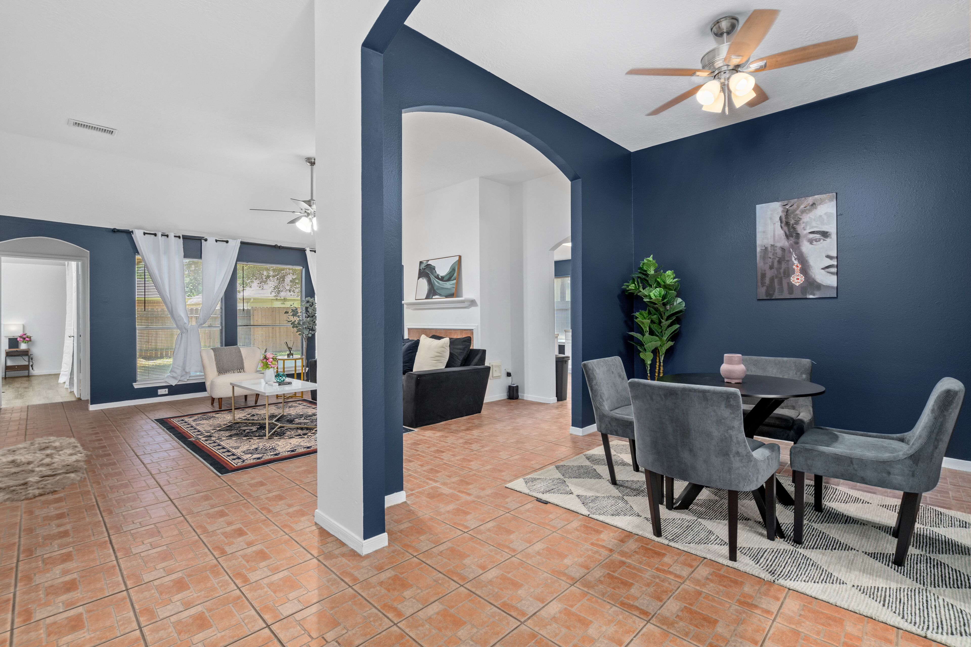 A spacious living area with a dining table, grey chairs, a ceiling fan, and a large window with curtains.