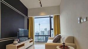 A modern living room with a large flat-screen TV, a beige sofa, a small round table with a decorative item, and a view of a cityscape through a large window. - The Axon Bukit Bintang by SMOVF BNB (Kuala Lumpur)