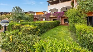 A well-maintained garden with hedges and a lawn, a building with balconies and outdoor seating, and a clear blue sky.