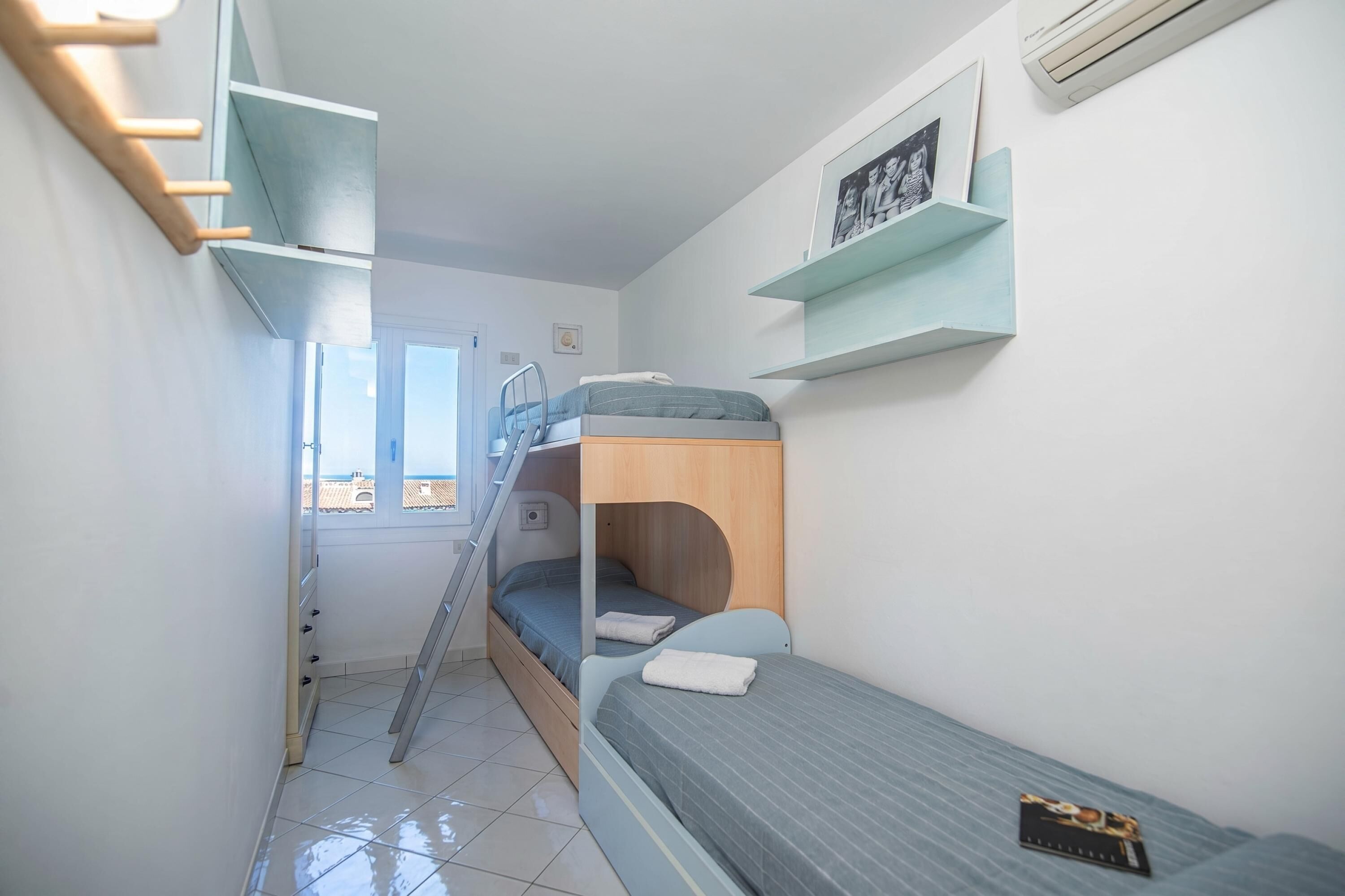 A compact room with a bunk bed, a ladder, a small window, and a wall-mounted shelf with a picture.
